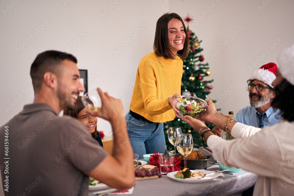 Beautiful young smiling Caucasian woman standing passing salad at Thanksgiving meal. Happiness at December festive vacation. Multi-generational people enjoying family reunion together Christmas. - obrazy, fototapety, plakaty 