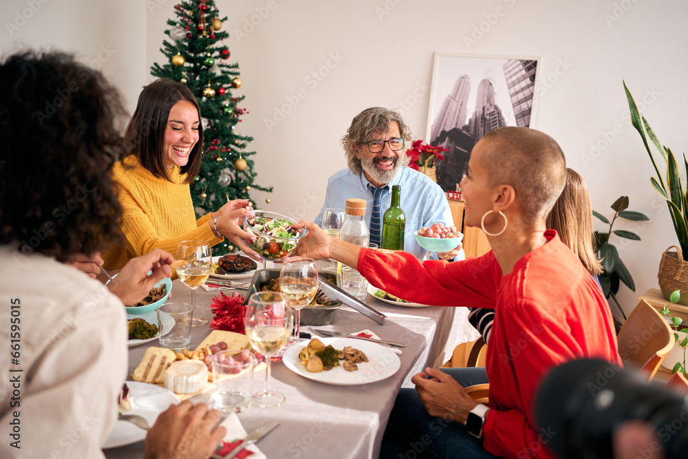 Happy family gathered around dining table during Christmas lunch at home. Women pass by and pick up salad bowl of smiling food. Domestic life in several generations and festive decoration with tree. Happy family gathered around dining table during Christmas lunch at home. Women pass by and pick up salad bowl of smiling food. Domestic life in several generations and festive decoration with tree.