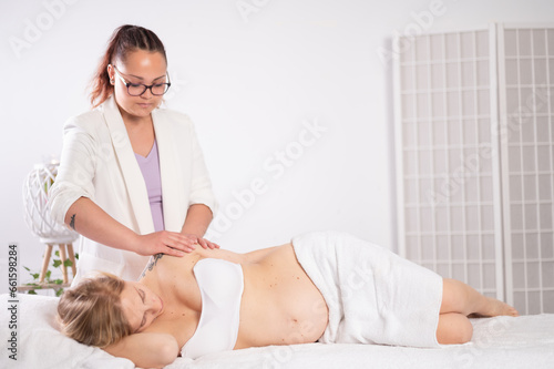 Ethnic masseuse massaging resting young pregnant woman on bed in parlor
