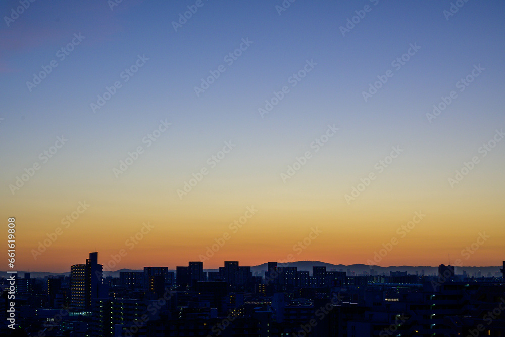 都市の夜明け。日の出とともに空と雲がオレンジ色に染まり、ビル群はシルエットとして写す。神戸市東灘区から大阪方面をのぞむ
