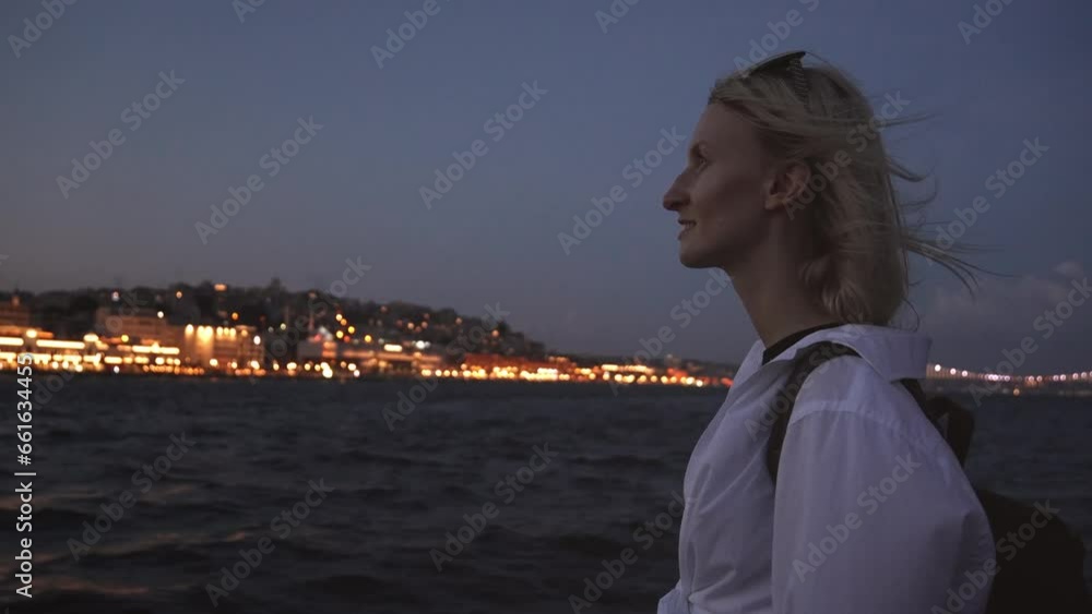 Young beautiful blonde woman in evening or at night on ferry Istanbul while relaxing Bosphorus. Girl in white stylish shirt on ship. Waves and city lights in background. Sightseeing boat around city Young beautiful blonde woman in evening or at night on ferry Istanbul while relaxing Bosphorus. Girl in white stylish shirt on ship. Waves and city lights in background. Sightseeing boat around city