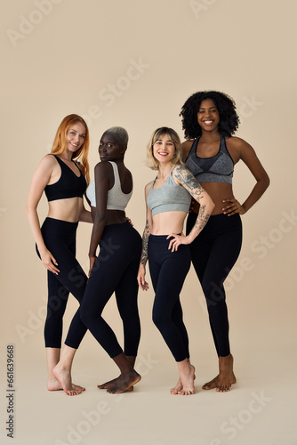 Happy fit sporty diverse young women wearing sportswear standing at beige background. Multicultural girls friends advertise sport fitness gym trainings together posing for vertical full body portrait.