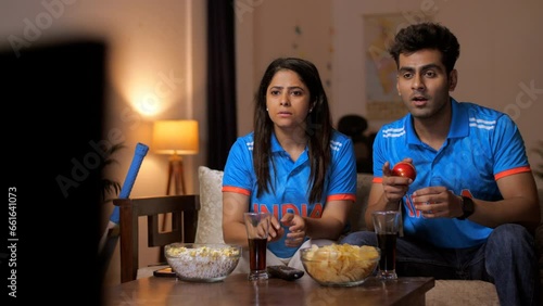 Friends in Indian jerseys sitting on the couch and watching a live cricket match on TV/ television - cricket fever, World Cup 2023. Indian cricket fan wearing an Indian jersey - excited  cheering 