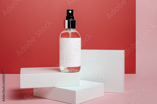 transparent spray bottle with silver cap and black spray top, blank white label, standing on white raisers on a red and pink background