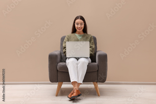 Beautiful woman with laptop sitting in armchair near beige wall indoors