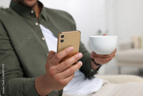 Man with cup of drink sending message via smartphone indoors, closeup