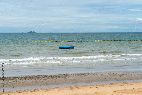 During low tide, fencing buoys and a blue platform float nearby in the tropical sea. Weak waves roll onto the sandy shore of the beach. The sky is overcast with light clouds.