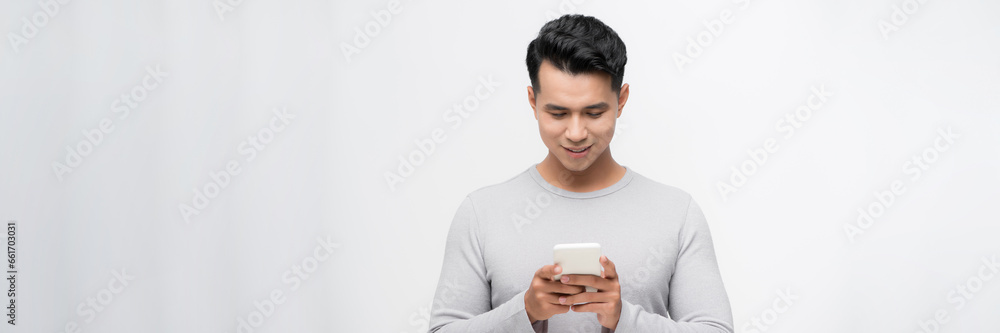 Fototapeta premium Handsome young man using his phone with smile while standing against white background.
