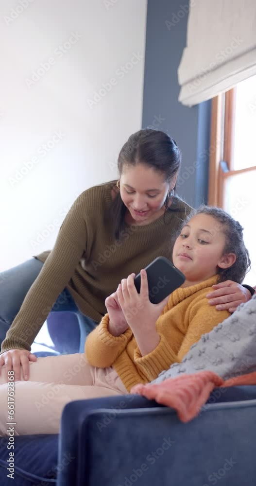 Happy biracial mother and daughter sitting on sofa and using smartphone in sunny living room Happy biracial mother and daughter sitting on sofa and using smartphone in sunny living room