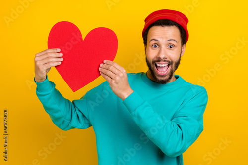 Portrait of ecstatic overjoyed man with beard dressed turquoise pullover demonstrate large paper heart isolated on yellow color background