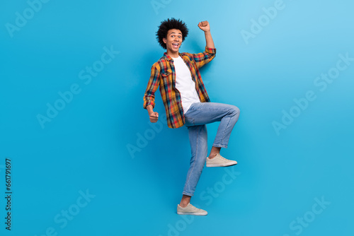 Full length photo of positive lucky guy wear plaid shirt walking rising fists screaming yeas isolated blue color background photo