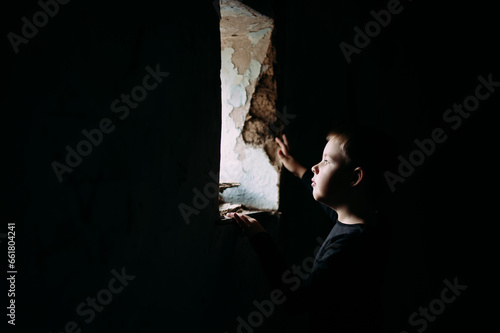 A little boy in a black T-shirt is standing in a house near a window. The child in the room is looking at the old window from which the light is coming. Consequences of war