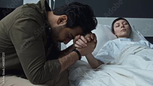 Selective focus medium close-up shot of worried Middle Eastern man holding hand of his beloved lying in hospital bed in coma photo