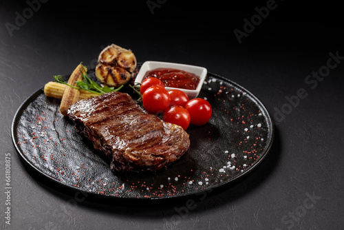 BBQ beef steak with sauce on a black plate with vegetables, tomatoes and corn. Black background. Restaurant supply.