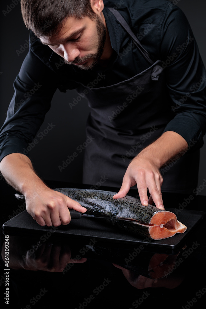 A cook on a black background cuts a whole salmon carcass. A cook on a black background cuts a whole salmon carcass.