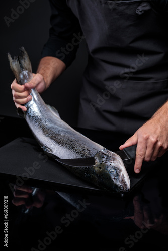 A cook on a black background cuts a whole salmon carcass.