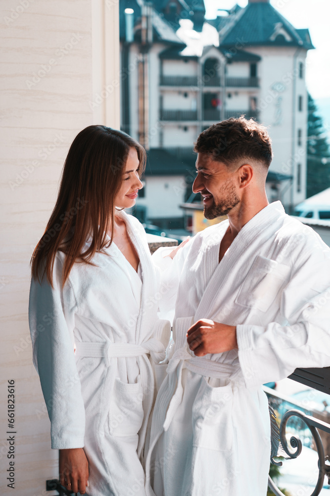 Close up of young couple in bathrobe standing on hotel balcony against mountain background - obrazy, fototapety, plakaty 