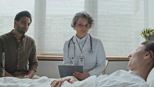 Medium shot of Caucasian female patient lying in bed explaining her symptoms to doctor while her Middle Eastern boyfriend attentively listening photo