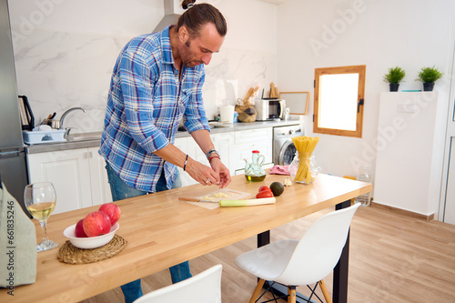Full length portrait of a young adult Caucasian handsome male chef in blue casual denim and plaid shirt, preparing dinner at home, chopping garlic on a cutting board. People. Lifestyle. Leisure