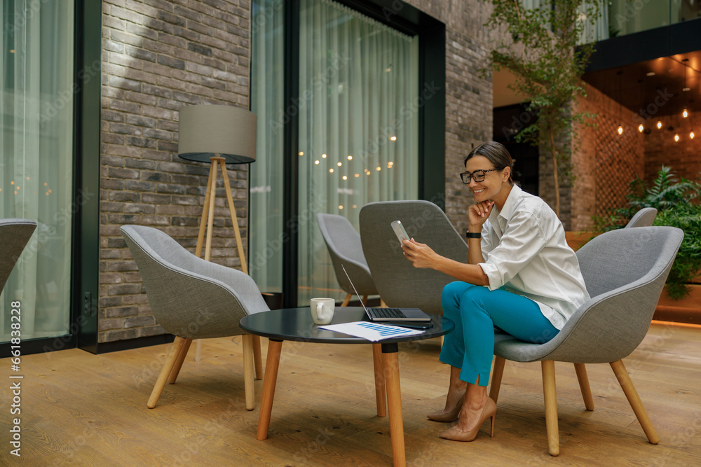 Pretty female entrepreneur in eyeglasses use phone while sitting in modern coworking space