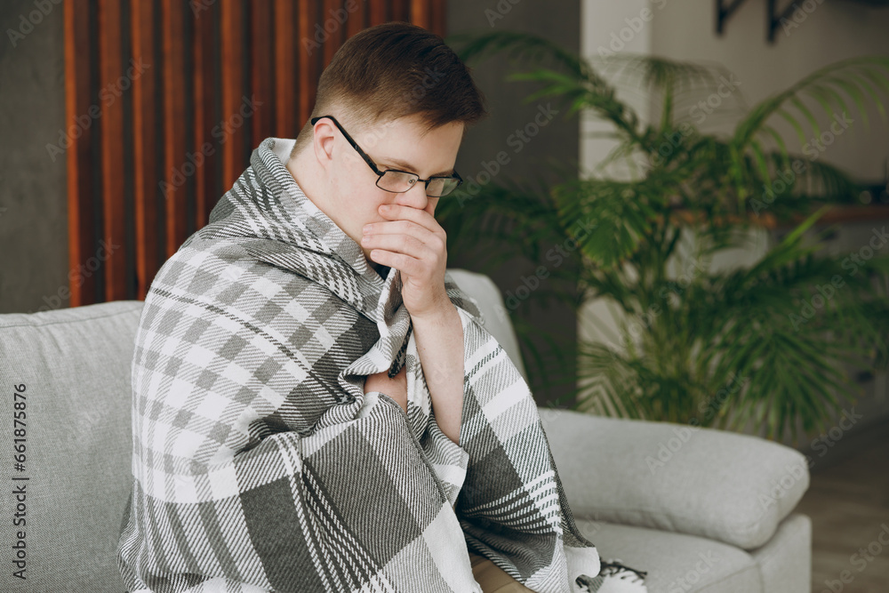 Poster Print Young sick ill tired exhausted man with down syndrome wears glasses wrapped in plaid sits on grey sofa couch stay at home flat rest