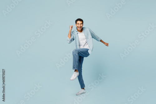 Full body young man of African American ethnicity he wear shirt casual clothes doing winner gesture celebrate clenching fists raise up leg isolated on plain pastel light blue cyan background studio.