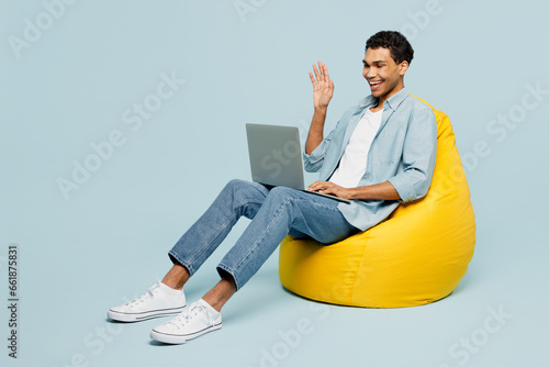 Full body young IT man of African American ethnicity he wear shirt casual clothes sit in bag chair hold use work on laptop pc computer waving hand isolated on plain pastel light blue cyan background.