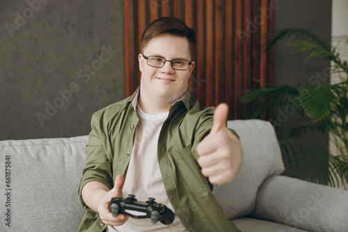 Young man with down syndrome wearing casual clothes play pc game with joystick console sits on grey sofa couch stay at home flat rest spend free time in living room. Genetic disease world day concept.