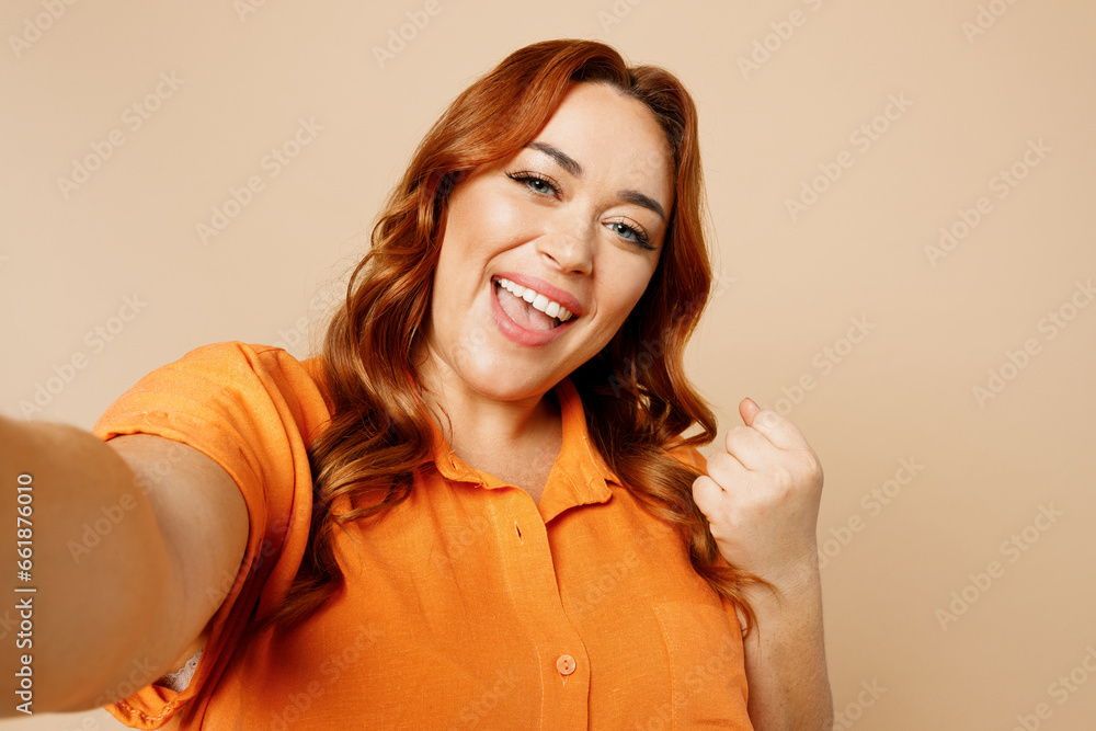 Close up happy young ginger chubby overweight woman wears orange shirt casual clothes doing selfie shot pov on mobile cell phone do winner gesture isolated on plain beige background studio portrait. Close up happy young ginger chubby overweight woman wears orange shirt casual clothes doing selfie shot pov on mobile cell phone do winner gesture isolated on plain beige background studio portrait.