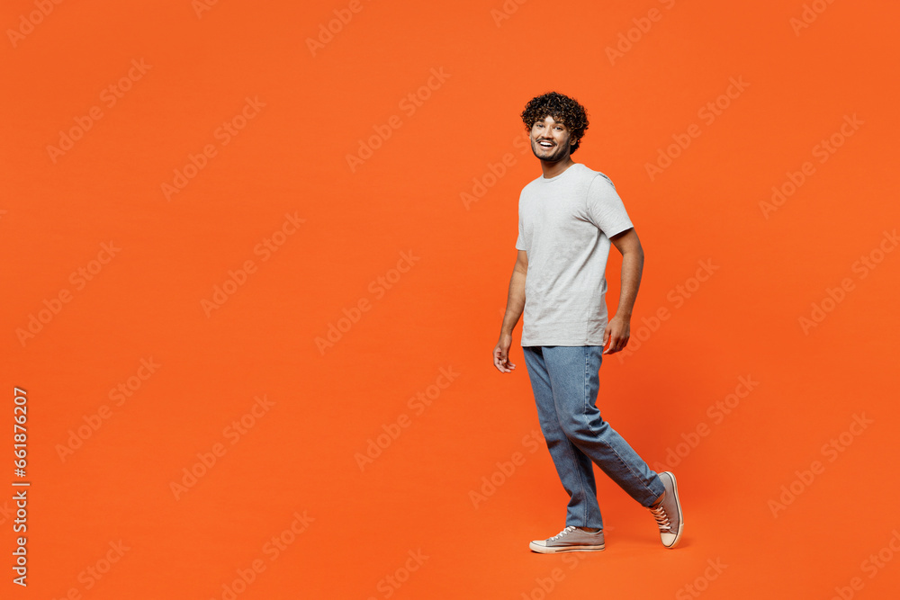 Full body side profile view young smiling happy Indian man he wearing t-shirt casual clothes walk going looking camera isolated on orange red color wall background studio portrait. Lifestyle concept. Full body side profile view young smiling happy Indian man he wearing t-shirt casual clothes walk going looking camera isolated on orange red color wall background studio portrait. Lifestyle concept.