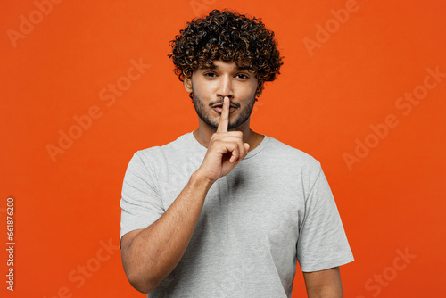 Young secret smiling fun happy Indian man he wears t-shirt casual clothes say hush be quiet with finger on lips shhh gesture isolated on orange red color background studio portrait. Lifestyle concept. photo