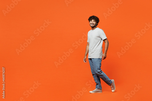 Full body side profile view young smiling happy Indian man he wearing t-shirt casual clothes walk going looking camera isolated on orange red color wall background studio portrait. Lifestyle concept.