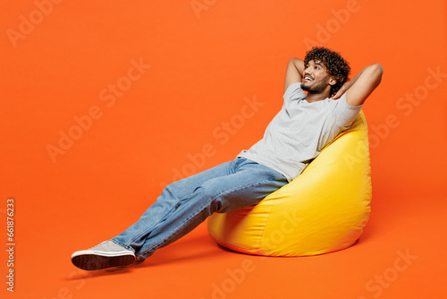 Full bodyside view young smiling happy Indian man he wear t-shirt casual clothes sit in bag chair hold hands behind head neck isolated on orange red color background studio portrait Lifestyle concept photo