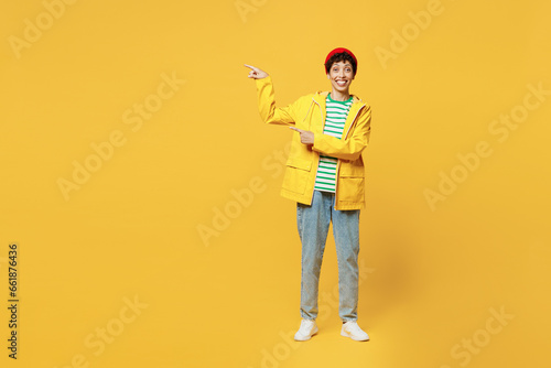 Full body young woman wearing waterproof raincoat outerwear red hat point finger aside on area isolated on plain yellow background studio portrait. Outdoors lifestyle wet fall weather season concept.