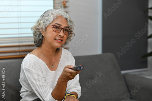 Senior Asian woman with remote control sitting on the sofa while watching television at home