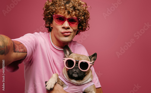 Cool young man holding French Bulldog pet in funky glasses and making selfie on pink background