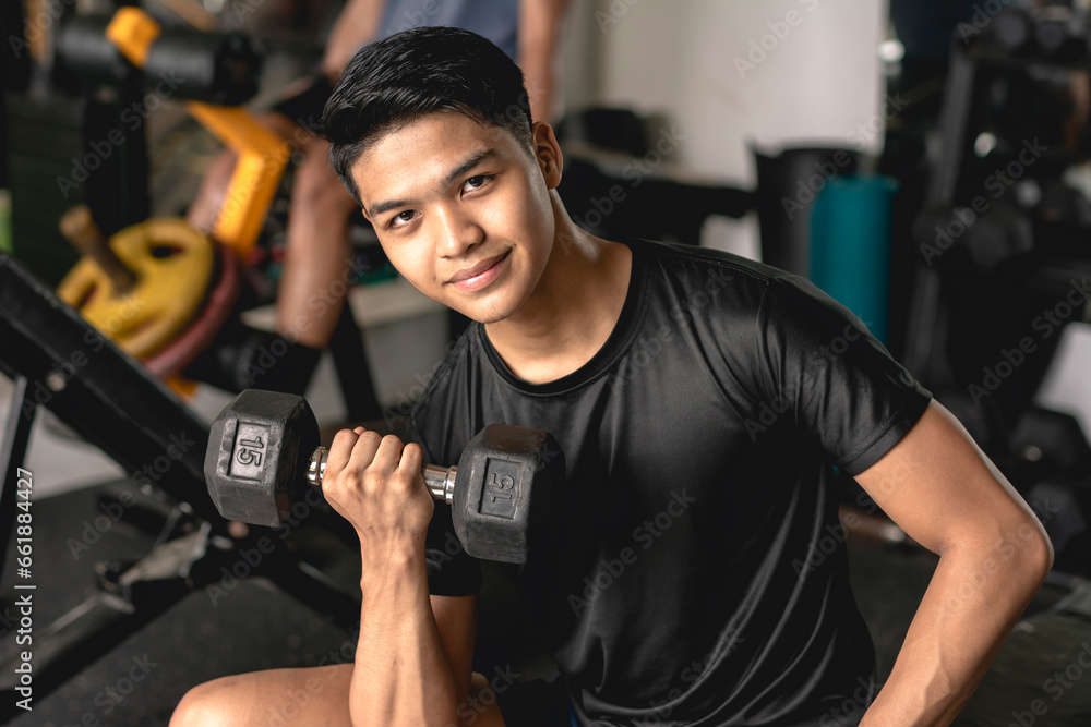 A young handsome asian man looking at the camera confidently while doing concentration bicep curls on the bench. Working out at the gym. A young handsome asian man looking at the camera confidently while doing concentration bicep curls on the bench. Working out at the gym.