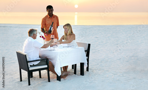 Couple, beach and dinner in sunset with waiter for romantic outdoor date, valentines day or anniversary. Man and woman with wine, cheers or celebration on table in relax or romance by ocean coast © Arcurs Corp/peopleimages.com