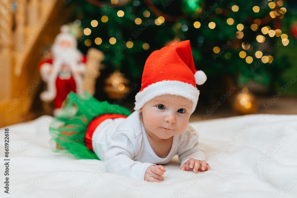 Baby girl with Christmas elf clothes is lying on a white blanket near Christmas tree with bokeh. Christmas concept. Infant baby in her first Christmas. Funny baby. Baby girl with Christmas elf clothes is lying on a white blanket near Christmas tree with bokeh. Christmas concept. Infant baby in her first Christmas. Funny baby.