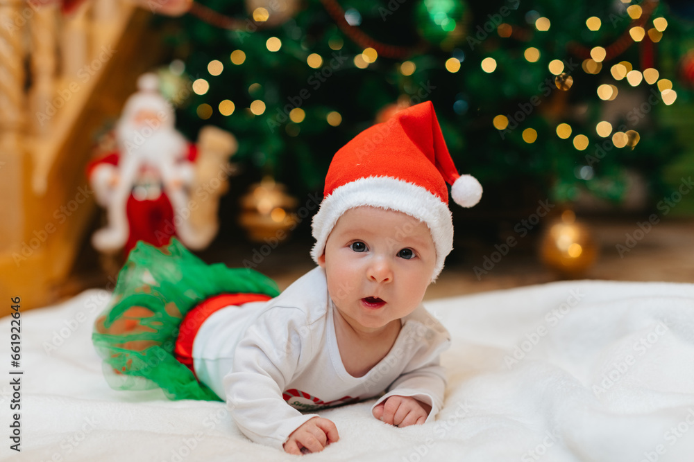 Baby girl with Christmas elf clothes is lying on a white blanket near Christmas tree with bokeh. Christmas concept. Infant baby in her first Christmas. Funny baby. Baby girl with Christmas elf clothes is lying on a white blanket near Christmas tree with bokeh. Christmas concept. Infant baby in her first Christmas. Funny baby.