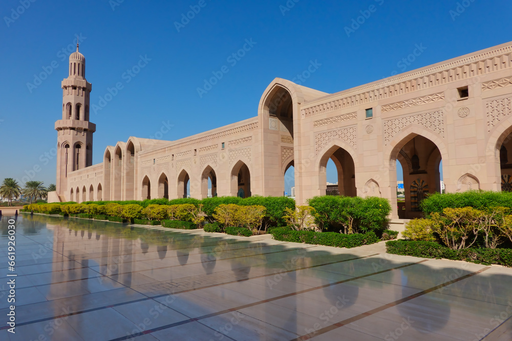 Fototapeta premium Exterior of the colonnade with the minaret of the Grand Mosque of Sultan Qaboos composed of many arches reflected on marble adorned with hedges and blue sky. Oman.