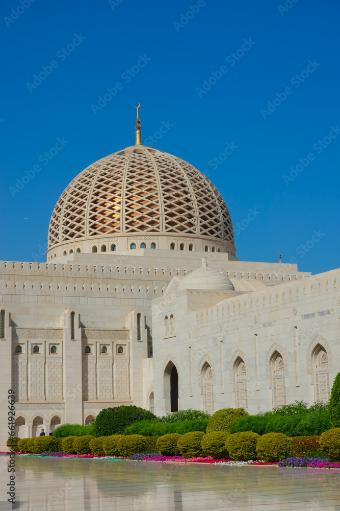 The exterior of the Grand Mosque of Sultan Qaboos in the foreground the dome decorated with geometric shapes adorned with gardens, in the background a blue sky. Oman.