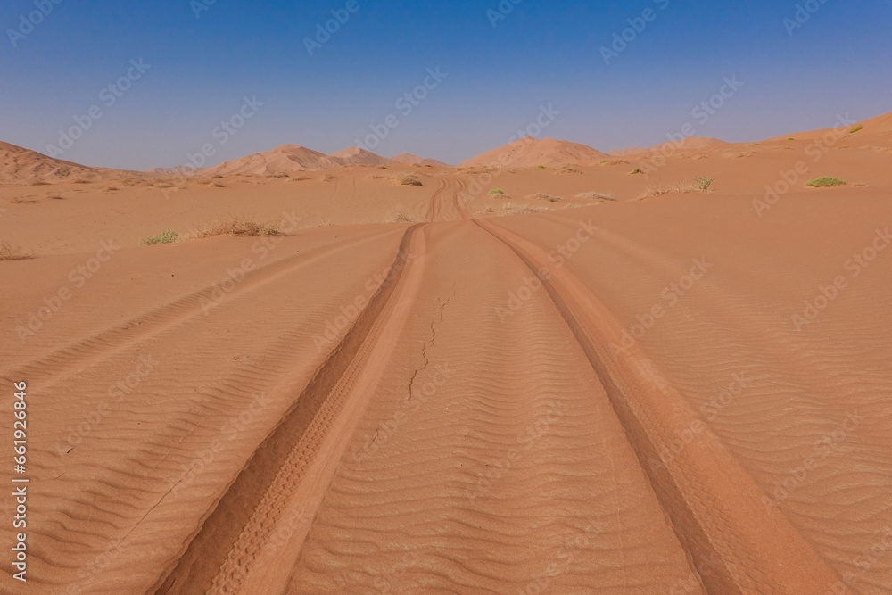 Tyre tracks on red sand in the Rub al Khali desert in the background the dunes and a blue sky. Oman.