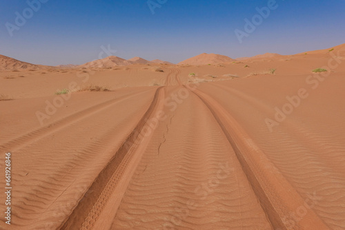 Tyre tracks on red sand in the Rub al Khali desert in the background the dunes and a blue sky. Oman.