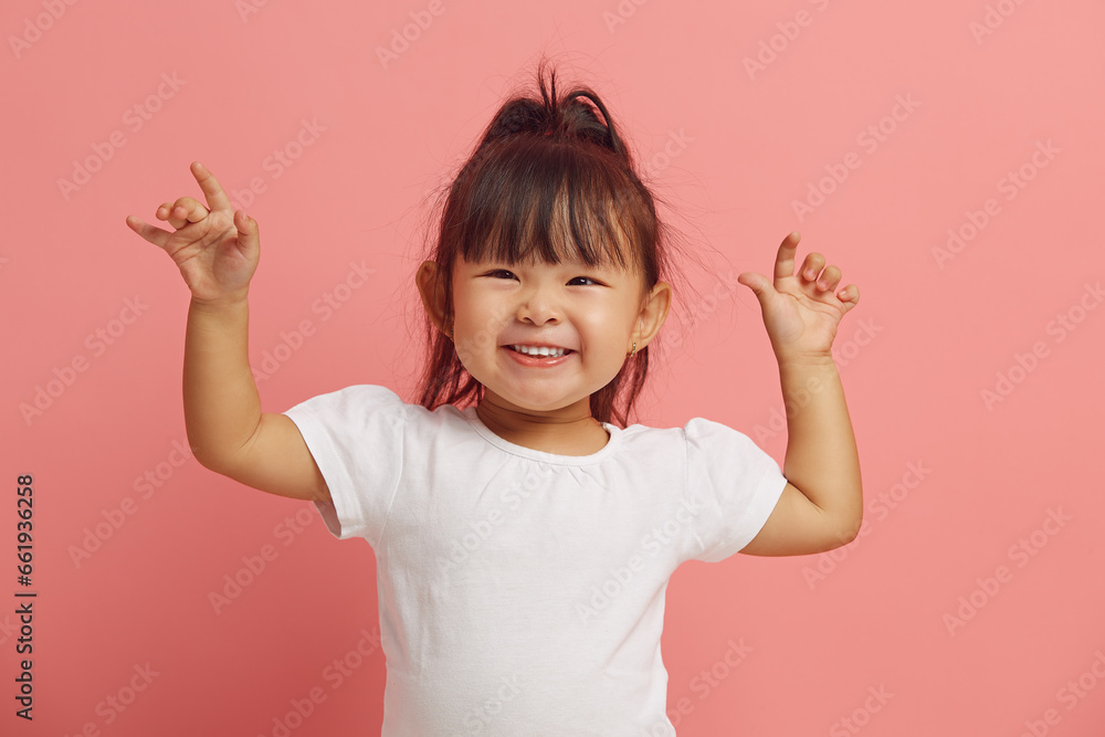 Joyful three years old Asian ethnicity little girl raises her hands up, expresses a cheerful mood, smiles happily standing on a pink isolated. - obrazy, fototapety, plakaty 