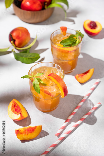 Two glasses with refreshing peach tea with ice and mint. Homemade cold summer drink on a light background with fresh fruits and shadows.