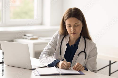 Female general practitioner in white coat sits at desk in hospital writes notes in paper notepad, work use laptop. Doctor manages appointment, fill patient anamnesis or client information in journal
