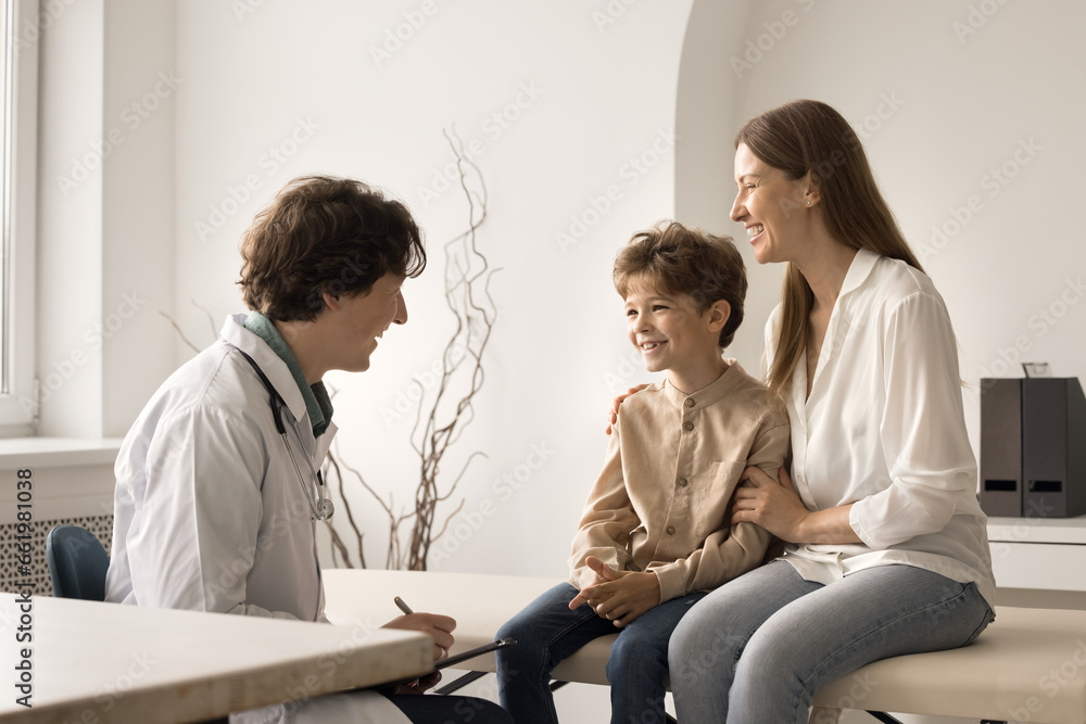 Obraz premium Little cute boy visit pediatrician doctor with loving mom. Friendly male paediatrician in coat hold clipboard writes symptoms, listen to kid patient, provide consultation. Healthcare, medical check-up