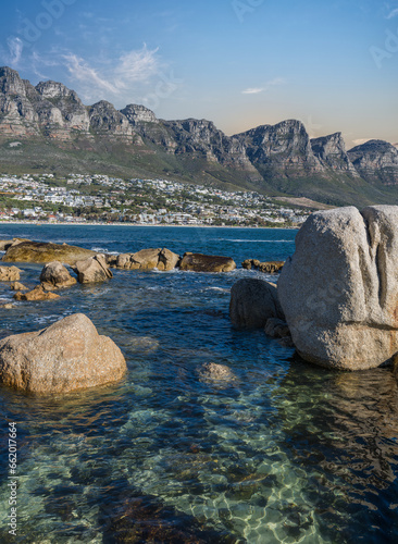 Shot of Camps bay town the Fourth Beach through the rocks and crystal clear atlantic ocean water ribbles during sunset with Twelve Apostles mountain range in the background, Cape Town, South Africa photo