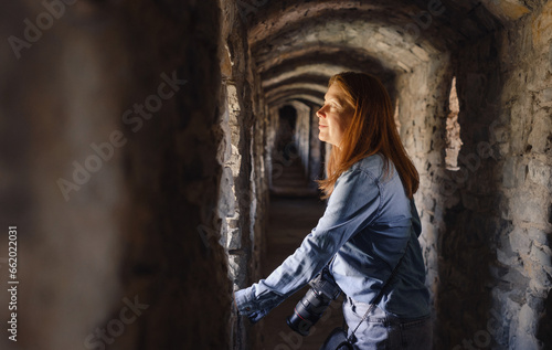 Fototapeta Naklejka Na Ścianę i Meble -  Woman walking through old tunnel, looking in window and taking pictures. Female visiting castle. Traveling in ancient building 